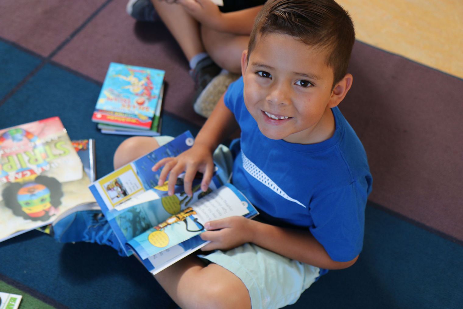 young boy smiling up at camera as he sits on the floor with a book