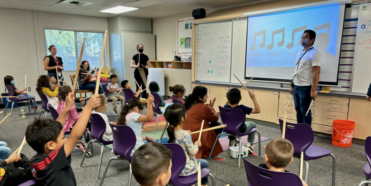male teacher standing at whiteboard with musical notes displayed as a dozen elementary boys and girls use drumsticks
