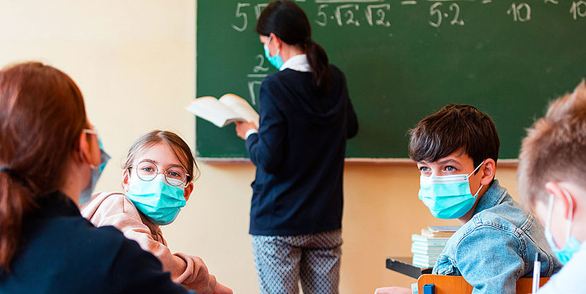 Teacher writing on the board and students talking wearing a mask.
