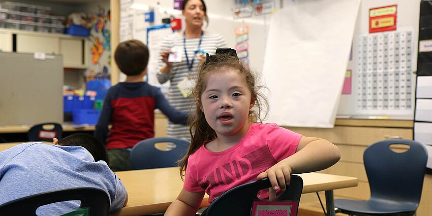 little girl with Down Syndrome in classroom looks at the camera as her teacher talks in the background.