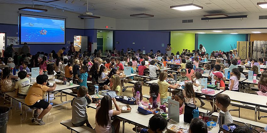 cafeteria filled with young students watching a video on large screen