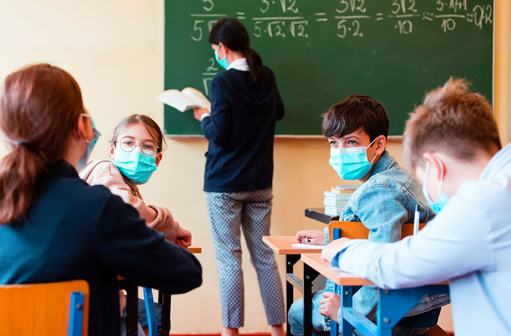 Teacher writing on the board and students talking wearing a mask.